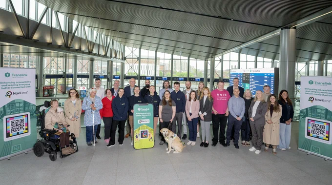 A group of people standing in an indoor space with a dog wearing a Guide Dogs harness. There are two large posters with the Translink logo.