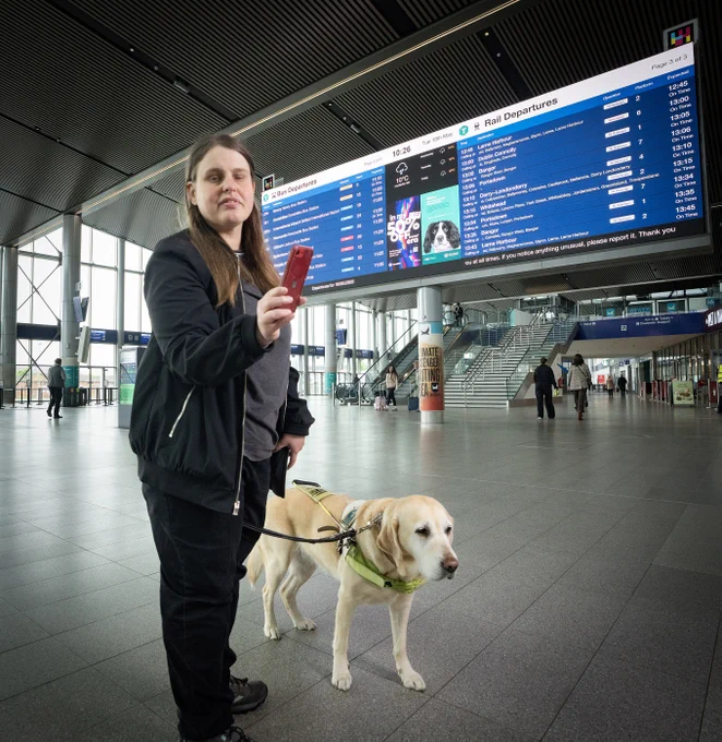 At Grand Central Station, a visually impaired woman with a guide dog checks hold a smartphone. Large displays list 'Bus Departures' and 'Rail Departures' with times.