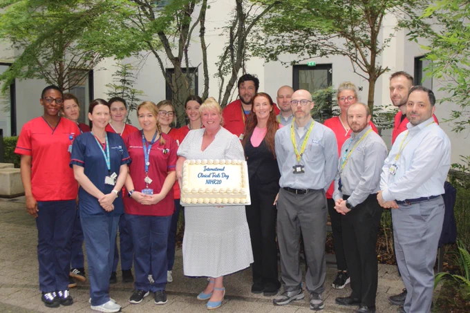A large group of colleagues from across Research and Innovation, standing together in one of the Royal Liverpool University Hospitals garden areas. The person in the middle of the Group is holding a large cake which has icing that reads 'International Clinical Trails Day 2026'.