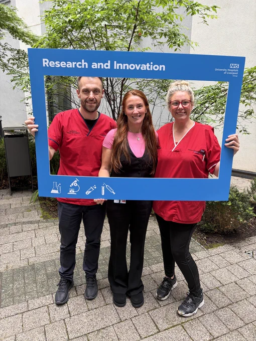 Three nurses in their uniforms holding a large blue picture frame which at the top reads 'Research and Innovation'. The nurses are smiling, stood in one of the Royal Liverpool University Hospitals garden areas.