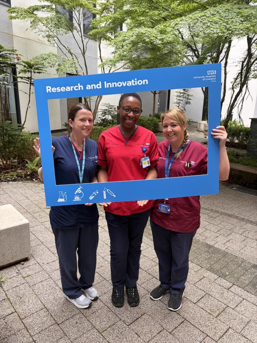 Three nurses in their uniforms holding a large blue picture frame which at the top reads 'Research and Innovation'. The nurses are smiling, stood in one of the Royal Liverpool University Hospitals garden areas.