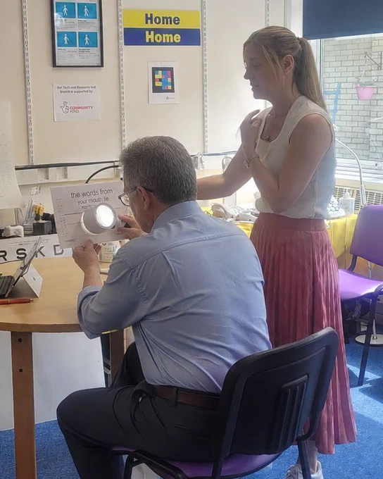 Woman holding an eye chart in front of a man sitting down looking through a magnifier.