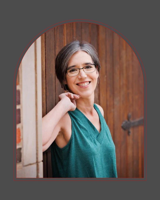 A white woman with black and gray hair in a short pixie cut. She's wearing glasses and a teal sleeveless shirt and leaning on a wide wooden door next to a brick wall.