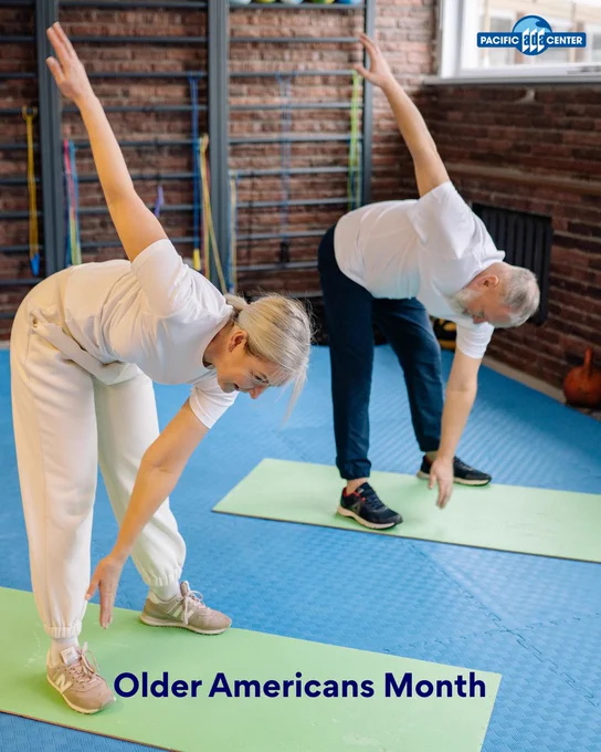 Two seniors performing side stretches on green mats in a fitness studio with brick walls and exercise bands it has blue text that reads Older Americans Month.
