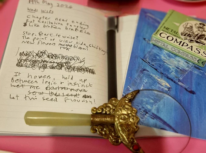 A close-up of a small open notebook showing handwritten text. The writing is messy, with errors crossed out using a fineliner pen with black ink. 
The text is a haiku which reads ‘chapter near ending, but hesitation tangles like broken branches. Stop. Start. Pause. Rewrite? The point of view side shifting –new flowers to bloom? It hovers, held up between logic and instinct – let this seed flourish!’ 
Where the notebook opens there is a thin black pen. To the right there is a blue postcard with an arty image showing blocks and vertical lines in subtle blue tones. On top in one corner is a small box reading ‘metal compass’, and below is an ornate magnifying glass with beautiful pewter embellishment. Everything is on a pink background.
