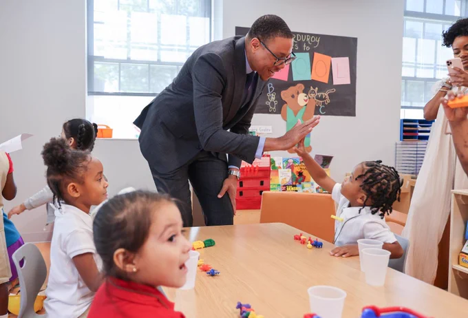 Chancellor Ferebee gives a high-five to a young DC student in a classroom. They are surrounded by other students playing with blocks sitting at a small desk.