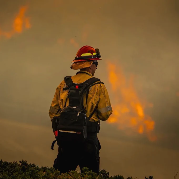 Fire Captain Jason Geiger of the huntington Beach Fire Department in protective gear, facing away, stands in front of a large wildfire. The sky is filled with smoke, and intense flames are visible in the background. The scene suggests active wildfire management. 
