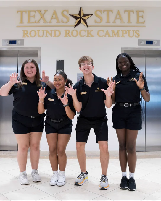 Four Texas State Round Rock Campus staff members in black uniforms posing with claw hand gestures in front of elevators.