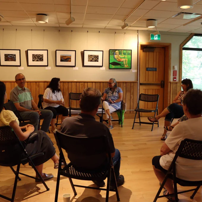 A group of people seated in a circle in a well-lit room, engaged in a discussion or meeting with framed artwork on the walls.