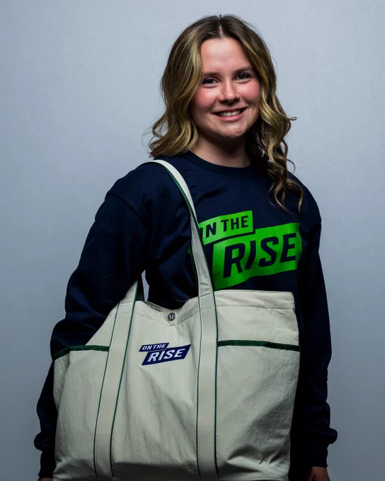 Photo of a women's student-athlete showing off the new OUA On the Rise Sweater and tote bag
