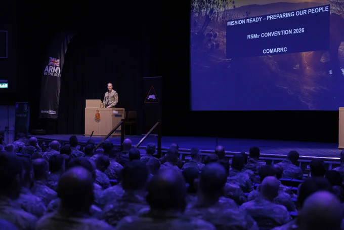 A person in military attire stands at a podium on a stage, addressing an audience of uniformed individuals. Large screens display the text "Mission Ready – Preparing Our People," "RSM’s Convention 2026," and "COMARRC." A British Army banner is visible to the left. The setting appears to be a formal military event in an auditorium.