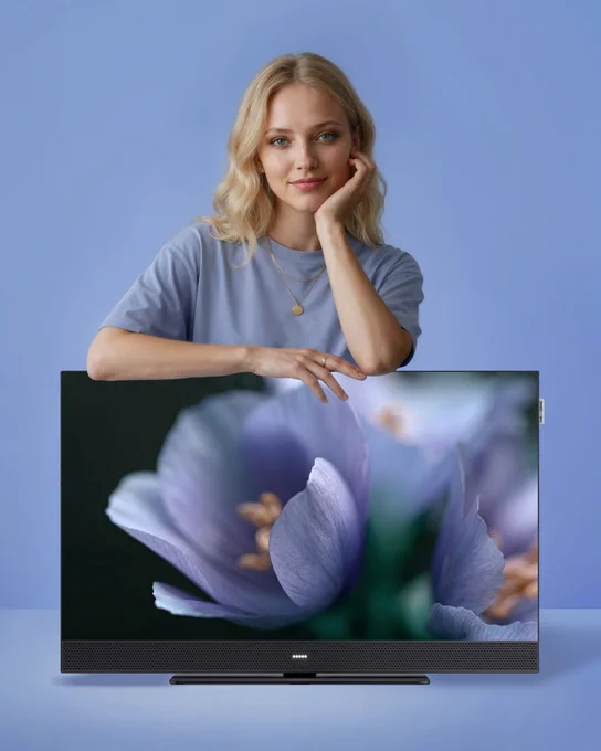 Blonde woman in a blue shirt leaning on a modern We. SEE lcd TV displaying a close-up of purple flowers against a blue background.