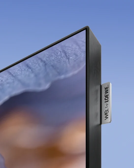 Close-up of a sleek, thin We. SEE lcd TV screen corner with a blurred nature image and an aluminium 'We by Loewe' label on the side.