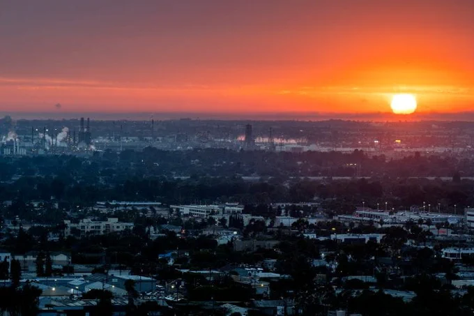 A vibrant sunset over an urban landscape with visible industrial structures and buildings. The sky is painted in shades of orange and red, casting a warm glow over the cityscape.