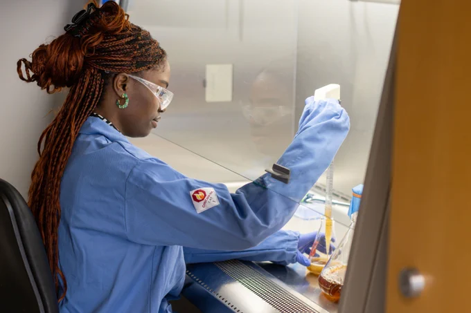 Damilola Olukorede is in protective glasses and a blue lab coat, conducting a laboratory experiment.