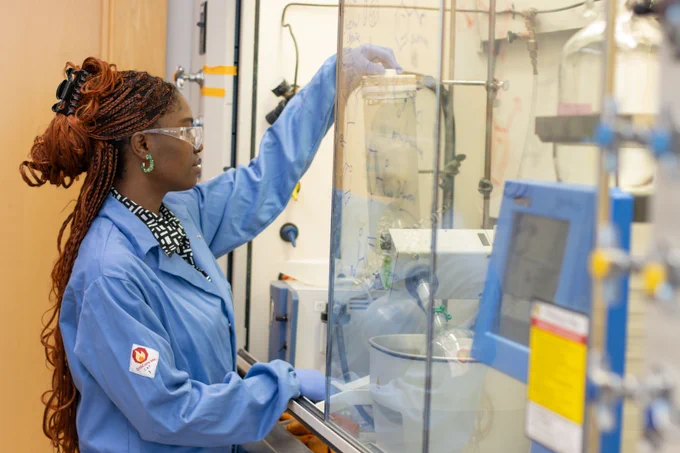 Damilola Olukorede in a lab coat and safety goggles, working at a chemistry lab station, adjusting equipment inside a fume hood.
