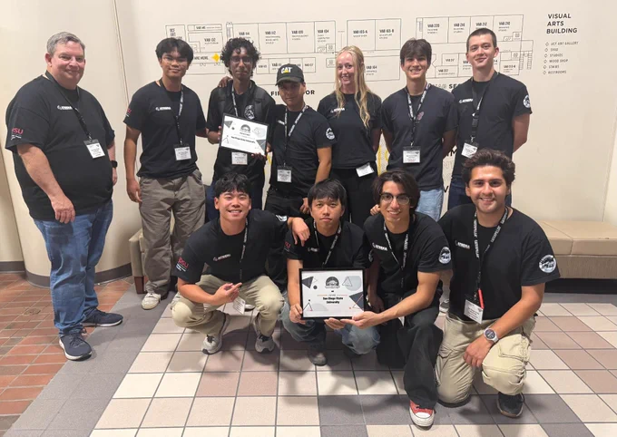 A group of individuals is posing together indoors, each wearing black shirts with various logos. Two people in the front are holding certificates. The background shows a wall with a detailed layout of a building.