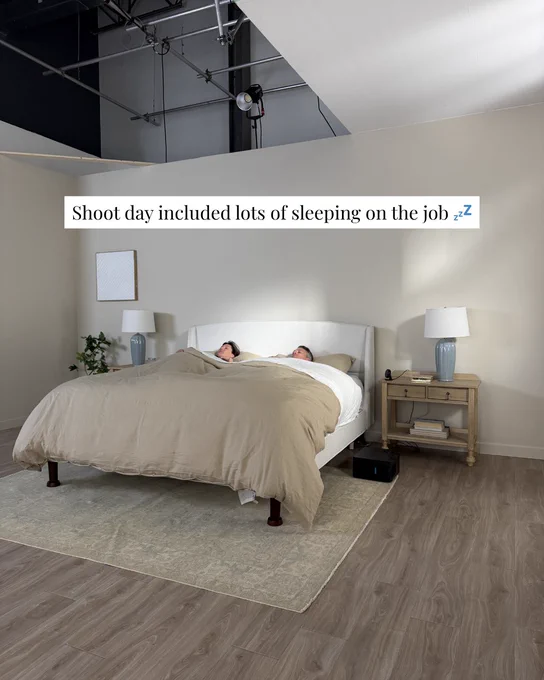 A staged bedroom scene with two people in bed during a photo shoot. The atmosphere is humorous and light-hearted.