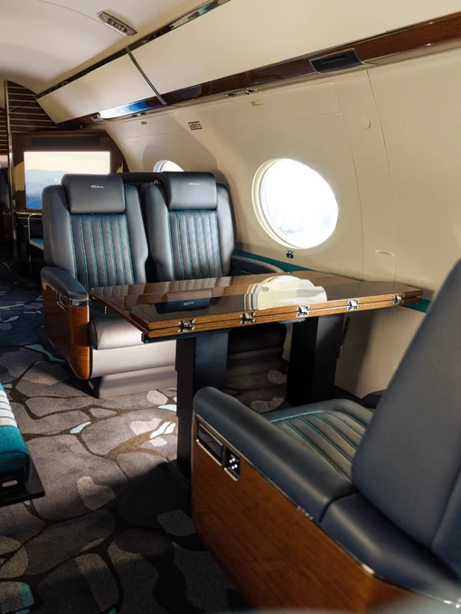 A luxurious interior of a private jet features leather seating and a polished wooden table. The cabin has oval windows and modern decor, providing a spacious and elegant atmosphere.