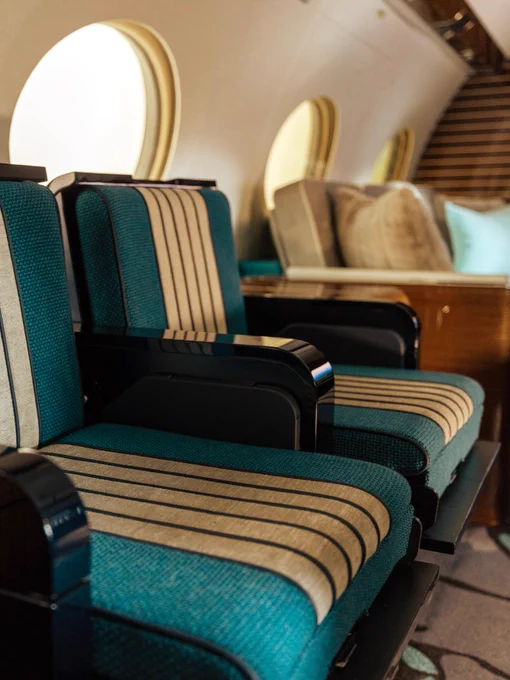 Luxurious seating inside a private jet cabin, featuring plush, striped teal and beige seats. The cabin has oval windows and elegant wood paneling. Pillows are arranged neatly on a side bench.