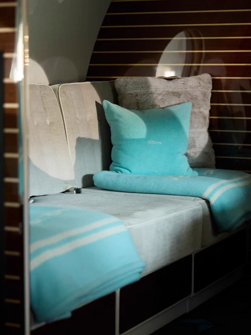 A cozy seating area inside a private space, featuring light-colored cushioned seats with a soft grey cushion and a blue pillow displaying "Riva." A matching blue blanket is draped elegantly across the seat. The background consists of a striped wooden panel.