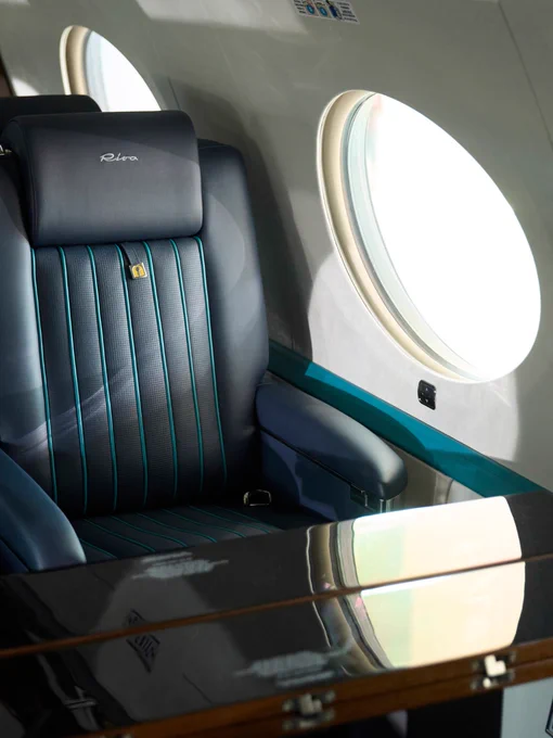 A luxurious airplane seat with dark upholstery and teal accents is positioned next to a round window. The headrest displays the logo "Pininfarina," and there is a table surface in front of the seat.