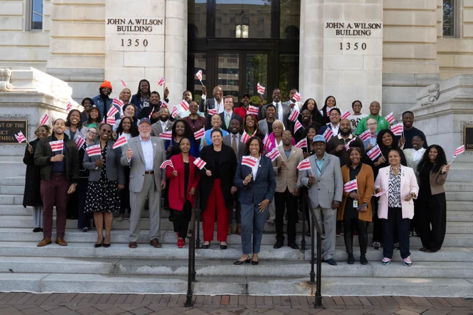 Mayor Bowser standing with a group of individuals on the front steps of the John A. Wilson Building. The group is holding DC flags.