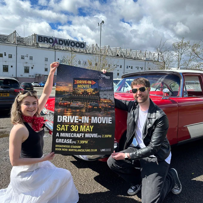 Two people dressed in retro attire hold a poster advertising a drive-in movie event at Broadwood Stadium next to a classic red car.
