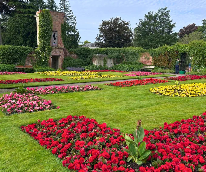 A formal garden with colourful flowerbeds of red, pink, and yellow flowers, set against a backdrop of hedges and a brick structure.