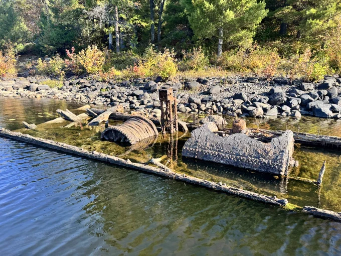 Weathered remains of an old alligator boat partially submerged along a rocky French River shoreline.