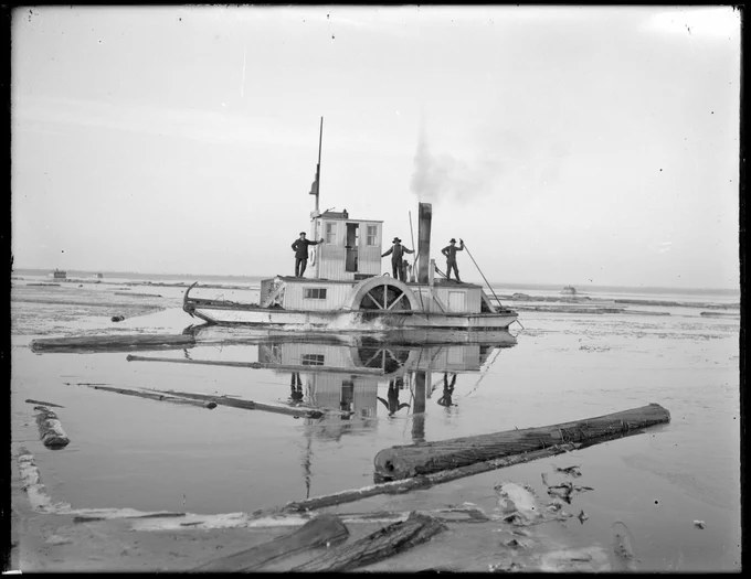Historic black‑and‑white photo of an alligator boat hauling logs through shallow water on the French River.