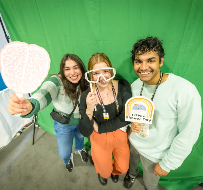 Three smiling individuals pose with playful brain-themed props against a green screen backdrop. They appear to be having fun, and the scene conveys a lighthearted and humorous atmosphere.