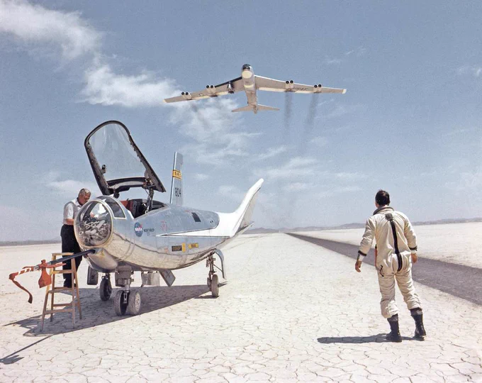 NASA research pilot Bill Dana takes a moment to watch NASA's NB-52B cruise overhead after a research flight in the HL-10. On the left, John Reeves can be seen at the cockpit of the lifting body. The dry lake bed at NASA Armstrong shines brightly in the sun.