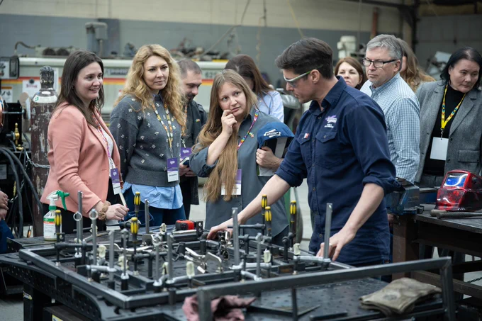 3 woman and other background participants observing a mechanical workshop, demonstrated by a male mechanic
