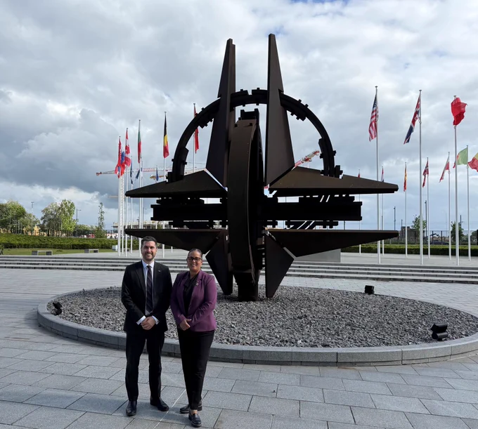 CSE’s Chief, Caroline Xavier, and CSIS Director, Dan Rogers, stand in front of the large NATO star sculpture surrounded by national flags.