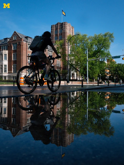 The Michigan Union reflected in a puddle as an individual rides a bike on State Street.