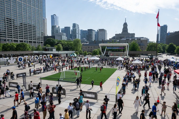 Crowd gathered around a small soccer field in a busy urban plaza with Toronto skyline and CN Tower in the background.