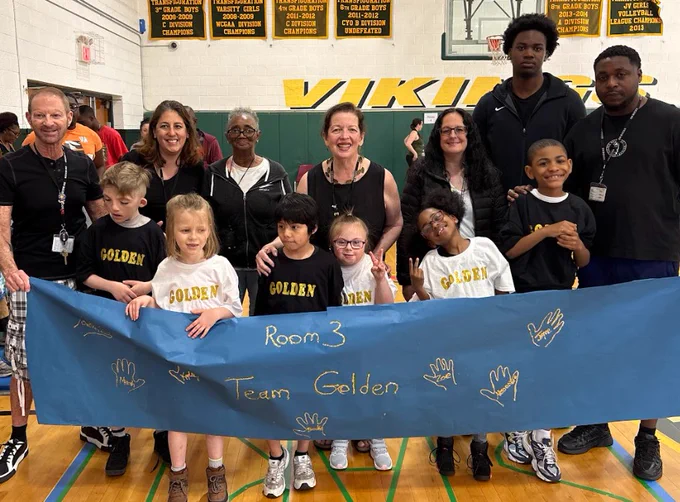 Group of children and adults in a gym holding a blue banner reading 'Room 3 Team Golden' with handprints and gold lettering.