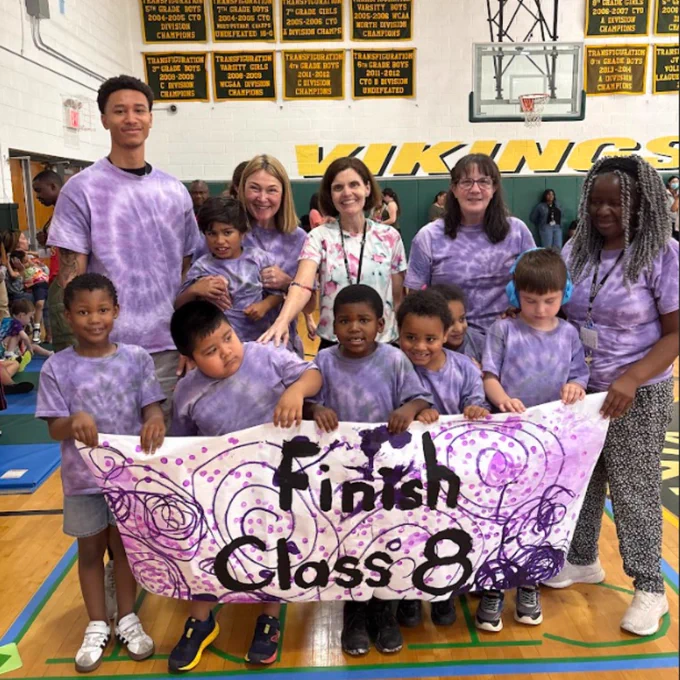 Group of children and adults in purple tie-dye shirts holding a decorated banner that reads 'Finish Class 8' inside a school gymnasium.