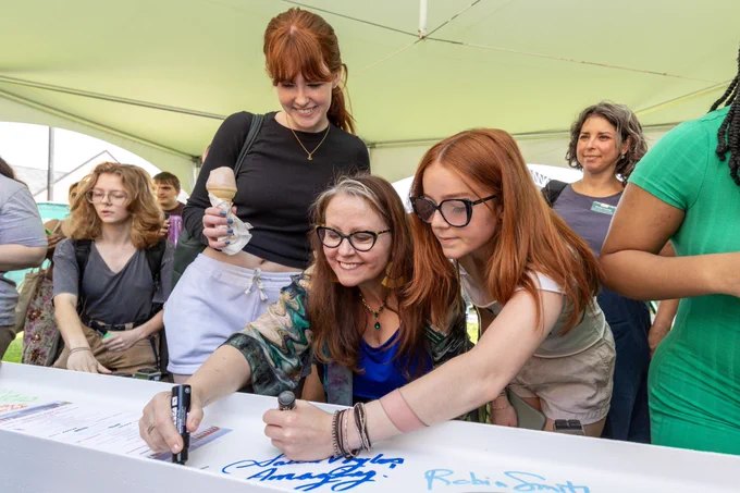 A group of people under a tent are gathered around a signing board. Several individuals are signing their names in large, colorful letters. Others are watching and smiling. The atmosphere appears friendly and engaging.