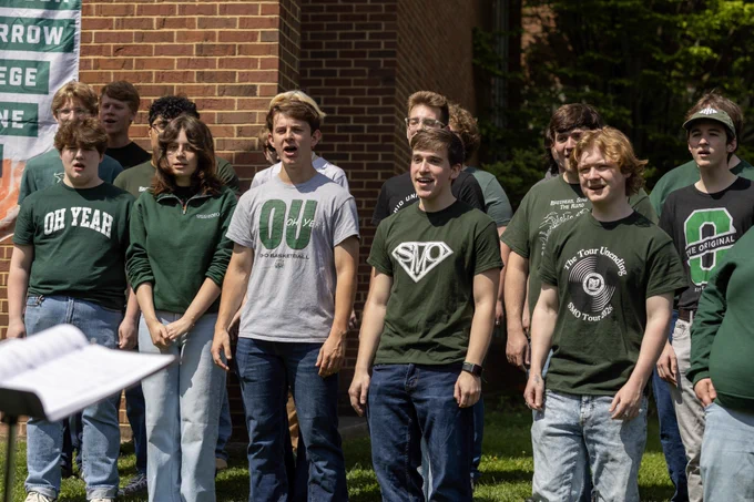 A group of people is singing outside near a brick wall. They are wearing casual outfits, primarily green shirts with various logos. A music stand with sheet music is visible in the foreground.