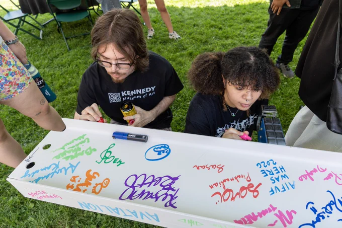 Two individuals are crouching on grass while writing colorful text on a large white beam. One is wearing a "Kennedy Museum of Art" t-shirt. Various names and doodles cover the beam. Other people are partially visible in the background.