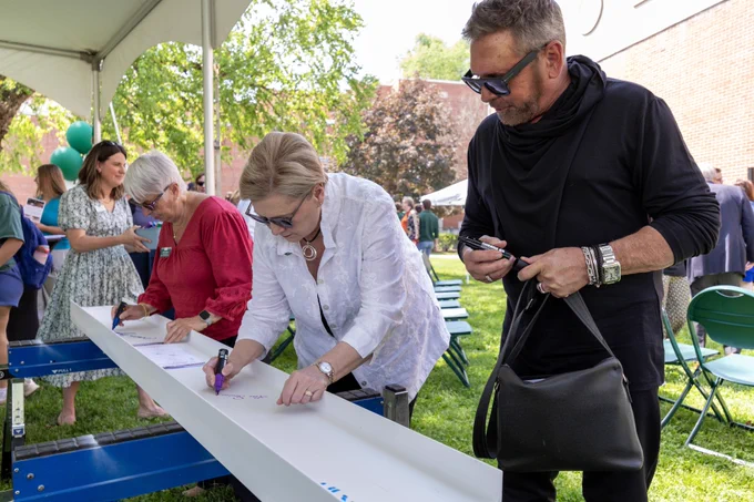 A group of people stand under a tent, signing a long, white beam positioned on a table, at an outdoor event. Trees and a brick building are visible in the background.