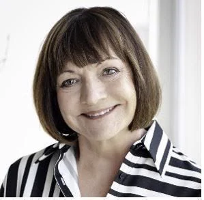 A headshot of Susan Kleimann smiling toward the camera. She has brown hair and bangs and is wearing a black and white striped shirt.