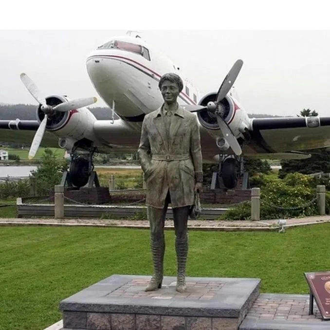 Amelia Earhart statue stands on a stone platform in front of a large twin-engine propeller airplane display on a manicured lawn. A small information plaque sits to the right on a low stone base, with trees, water, houses, and hills in the background under an overcast sky.
