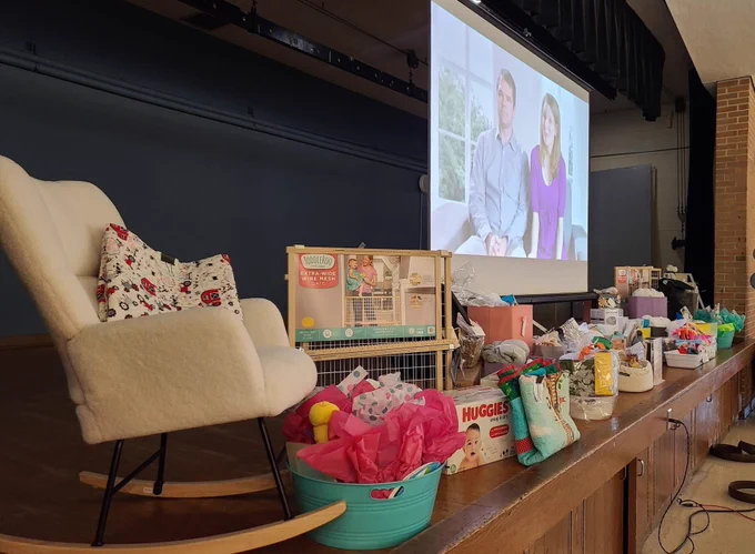 A stage is set up for an event, featuring a large screen. An array of gift items, including diapers, toys, and colorful baskets, are arranged on the stage. A cozy rocking chair and balloon decoration are also present.