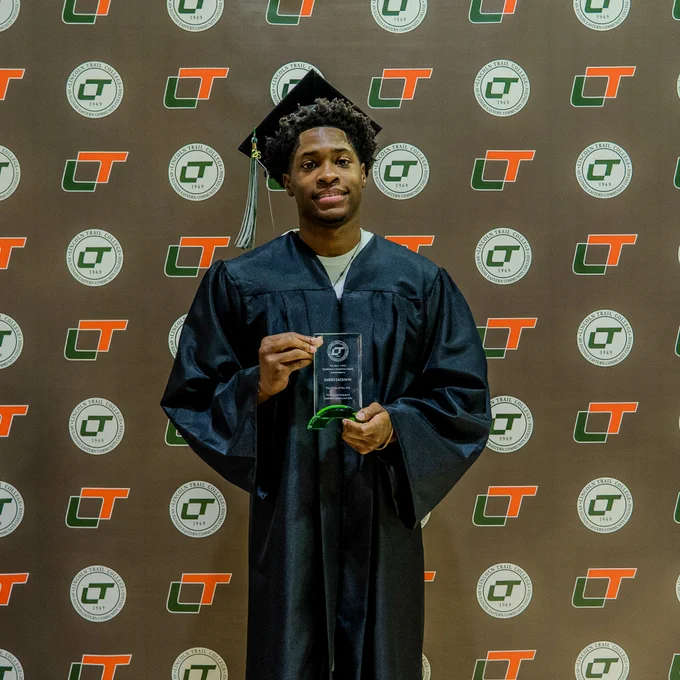 Jared Jackson poses in his graduation gown while holding the Bev L. Turkal Excellence in Leadership Award during Lincoln Trail College Commencement. He stands in front of an LTC backdrop featuring the college logo.