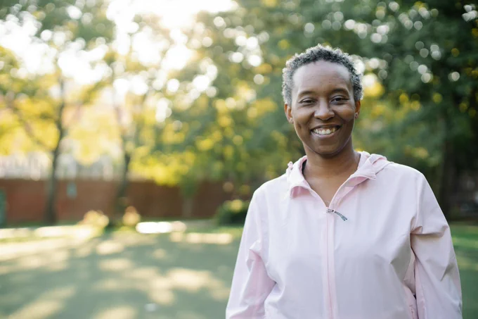 A portrait of an individual smiling outdoors with greenery and sunlight in the background.