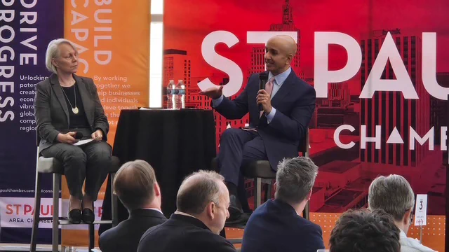Two individuals are seated on a stage, engaged in a discussion. One holds a microphone and gestures with a hand while speaking. Behind them are banners for the "St. Paul Area Chamber." Several audience members are visible in the foreground.