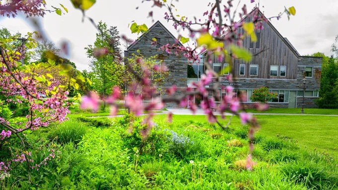 A scenic view of the Joel and Elizabeth Johnson Center for Health & Wellness is surrounded by vibrant green grass and blossoming trees. The foreground features branches with pink flowers, adding color to the natural setting.